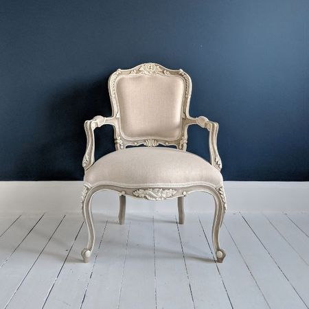 Elegant vintage armchair with ornate carvings and beige upholstery against a dark blue wall on a white wooden floor.