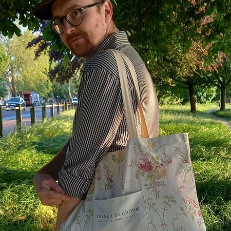 Man in glasses and hat holds a floral tote bag on a sunny path lined with trees and grass.