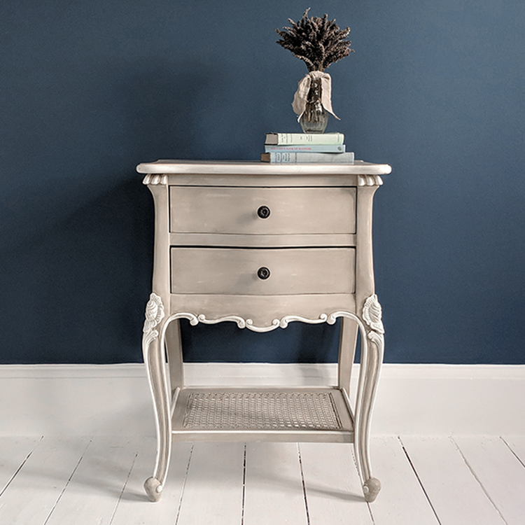 Elegant gray nightstand with two drawers, ornate legs, and a wicker shelf, topped with books and a dried flower bouquet against a blue wall.