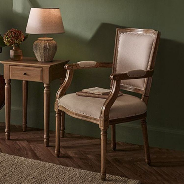 A vintage wooden armchair with beige upholstery next to a wooden side table holding a lamp and a small plant, set against a green wall.
