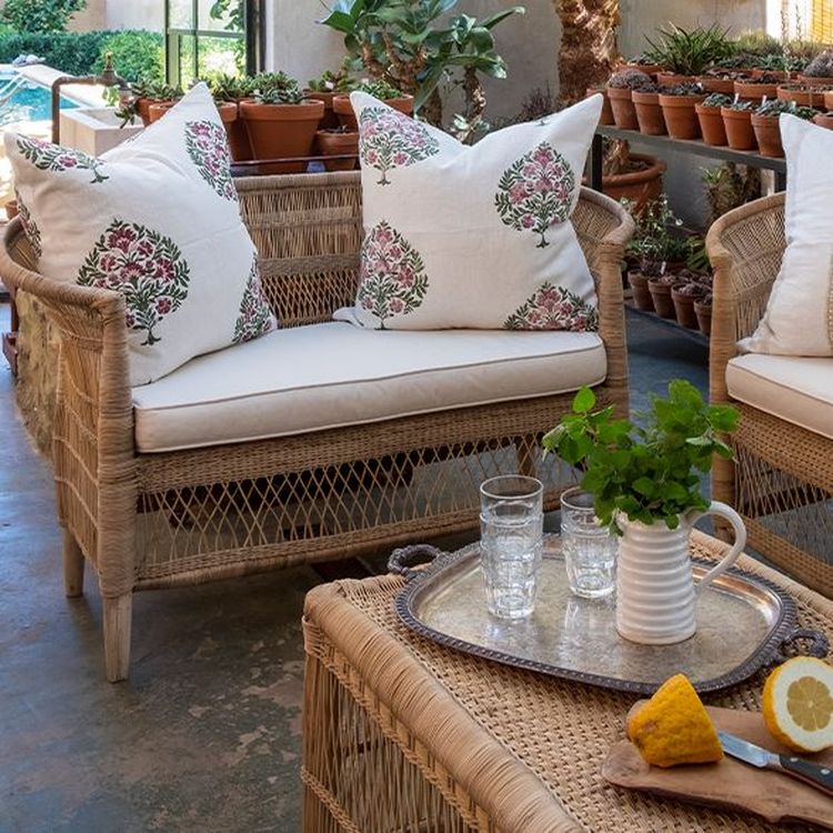 Wicker seating with floral cushions on a patio, surrounded by potted plants. A tray with glasses, a jug, and sliced lemon is on a wicker table.