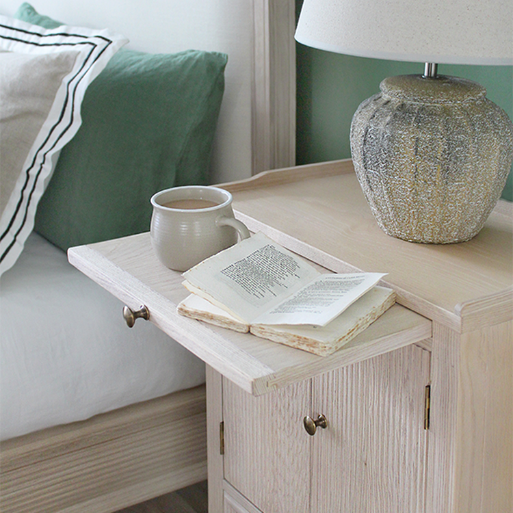 Cozy bedroom scene with a wooden nightstand, a lamp, an open book, and a mug. A bed with pillows is partially visible.