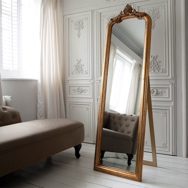 Elegant room with a tall, ornate gold-framed mirror reflecting a tufted chair, white paneled walls, and sheer curtains.
