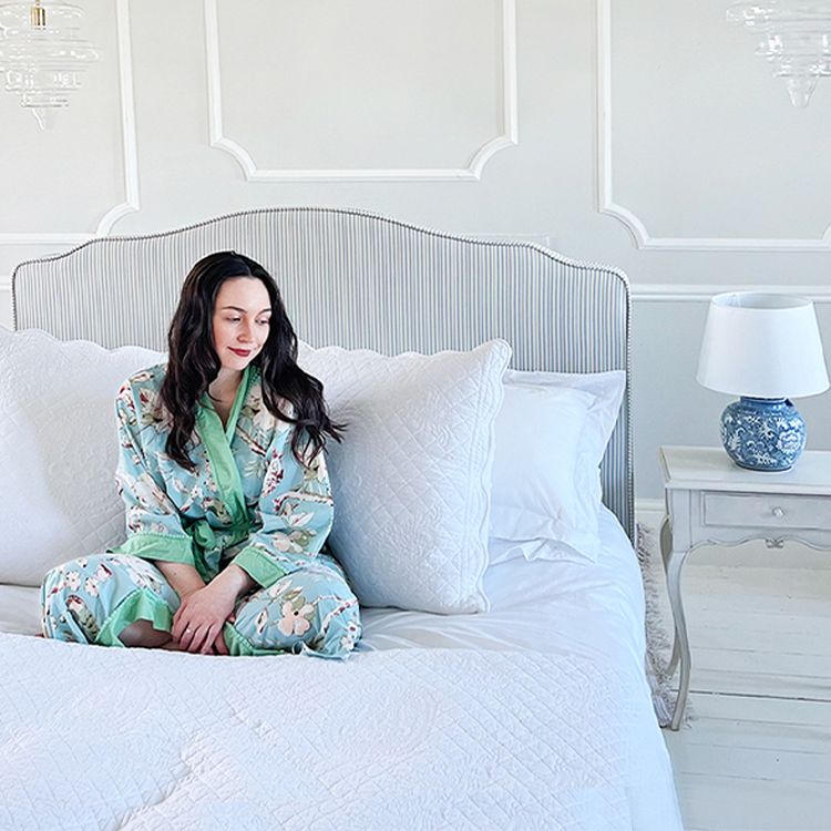 Woman in floral robe sits on a neatly made bed with white linens, next to a bedside table with a blue and white lamp, in a bright room.