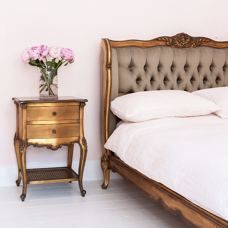 Elegant bedroom with a tufted headboard, ornate wooden nightstand, and a vase of pink flowers, all against a soft pink wall.