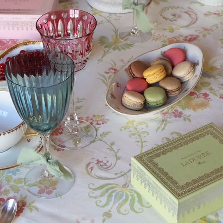 A table set with colorful macarons, elegant glassware, and a Ladurée box on a floral tablecloth.