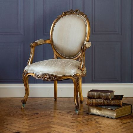 Ornate vintage chair with gold frame and beige upholstery beside a stack of old books on a wooden floor against a paneled wall.