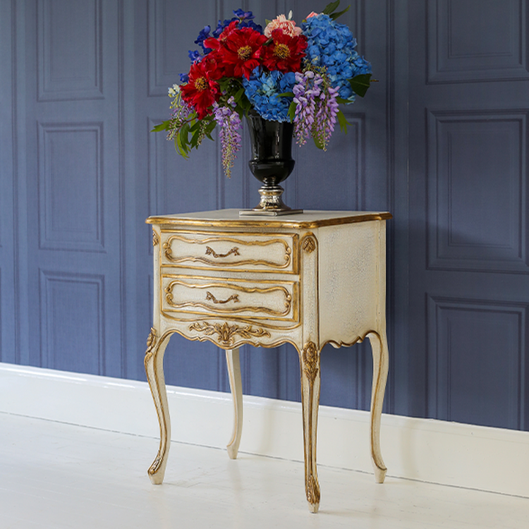 Elegant vintage side table with ornate details, topped with a black vase of vibrant red, blue, and purple flowers against a blue paneled wall.