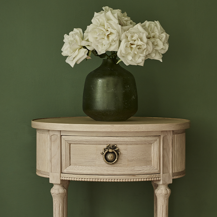 A round wooden table with a drawer, featuring a green vase holding white roses, set against a green wall.