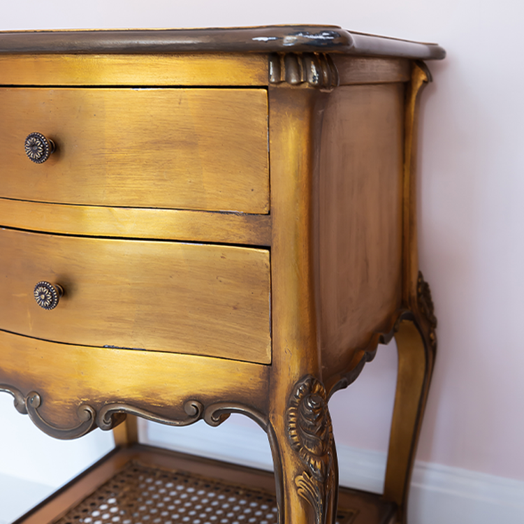 Golden vintage wooden dresser with ornate carvings, two drawers with round knobs, and a lower shelf. Set against a light pink wall.