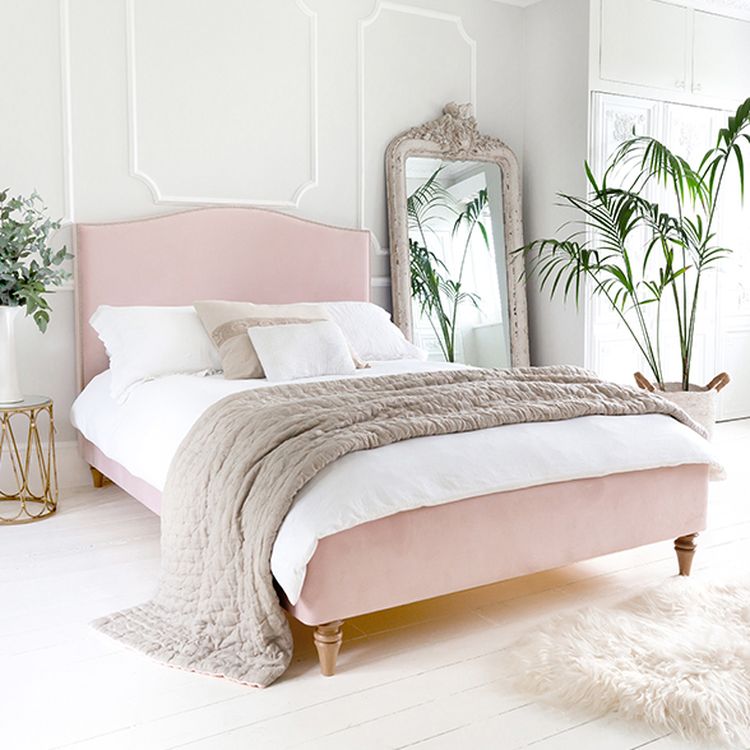 Elegant bedroom with a pink bed, white bedding, beige throw, large ornate mirror, palm plant, and a fluffy rug on white wooden floor.