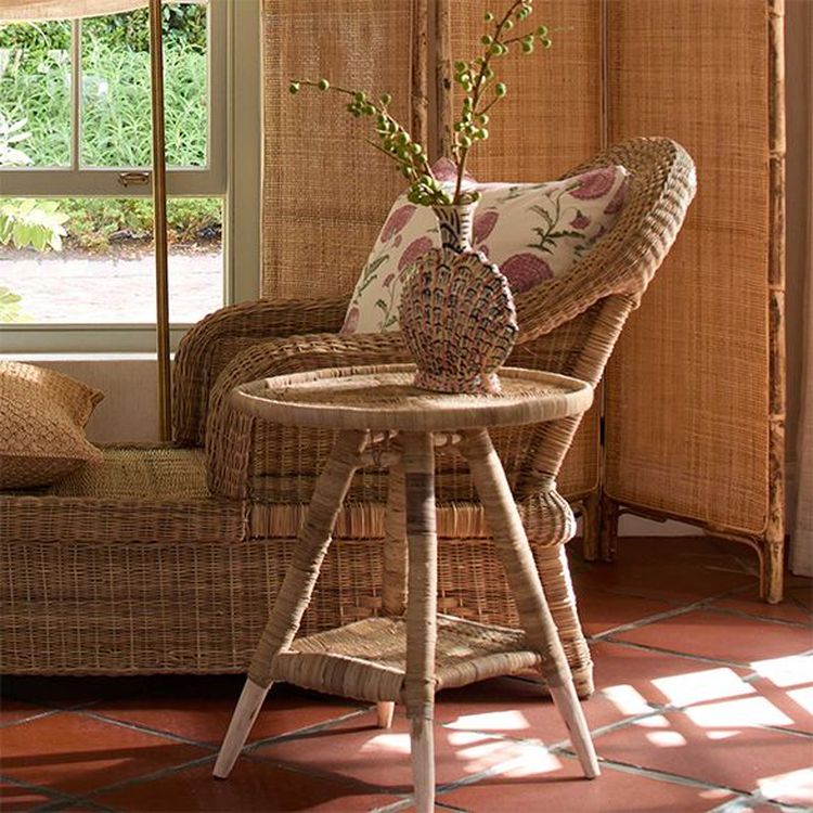 Cozy wicker chair with floral cushion beside a matching side table holding a vase of greenery, set on a terracotta tiled floor by a window.
