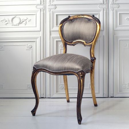 Elegant vintage chair with a gold-trimmed wooden frame and gray upholstered seat, set against an ornate white paneled background.