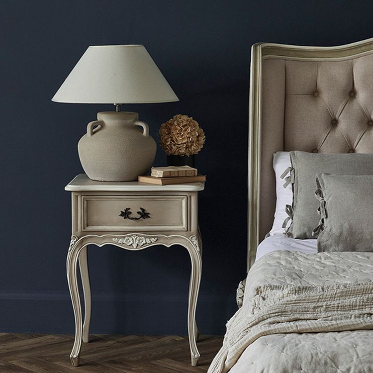 Elegant bedroom with a vintage nightstand, beige lamp, books, and dried flowers beside a tufted headboard and textured bedding on a wooden floor.