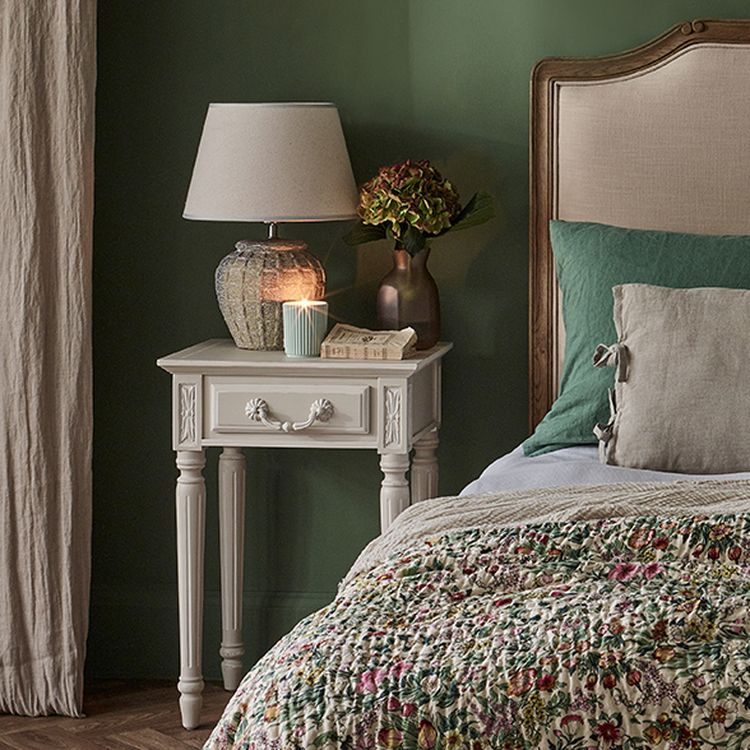 Cozy bedroom with a floral quilt, green pillow, and beige headboard. A white nightstand holds a lamp, candle, and flowers against a green wall.