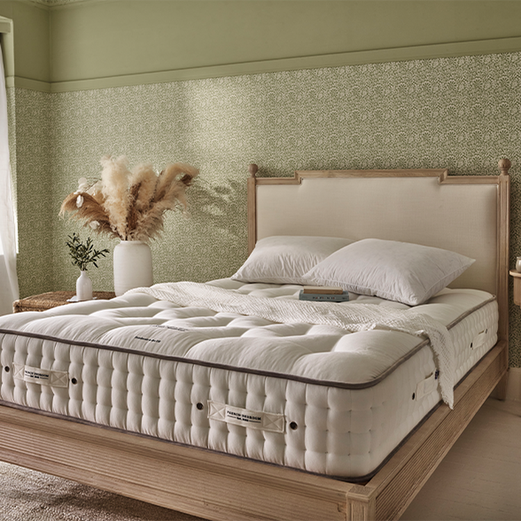 Cozy bedroom with a wooden bed frame, white mattress, pillows, and a vase of pampas grass. Light green patterned wallpaper adds a soft touch.