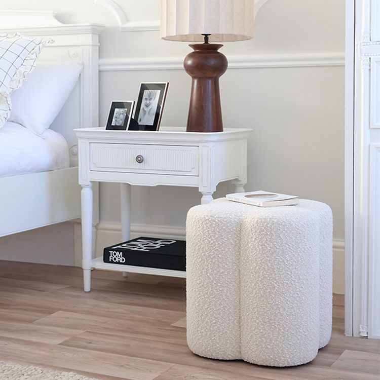 Bedroom corner with a white nightstand, brown lamp, framed photos, and a plush white ottoman on wooden flooring. A book titled "Tom Ford" is beneath.