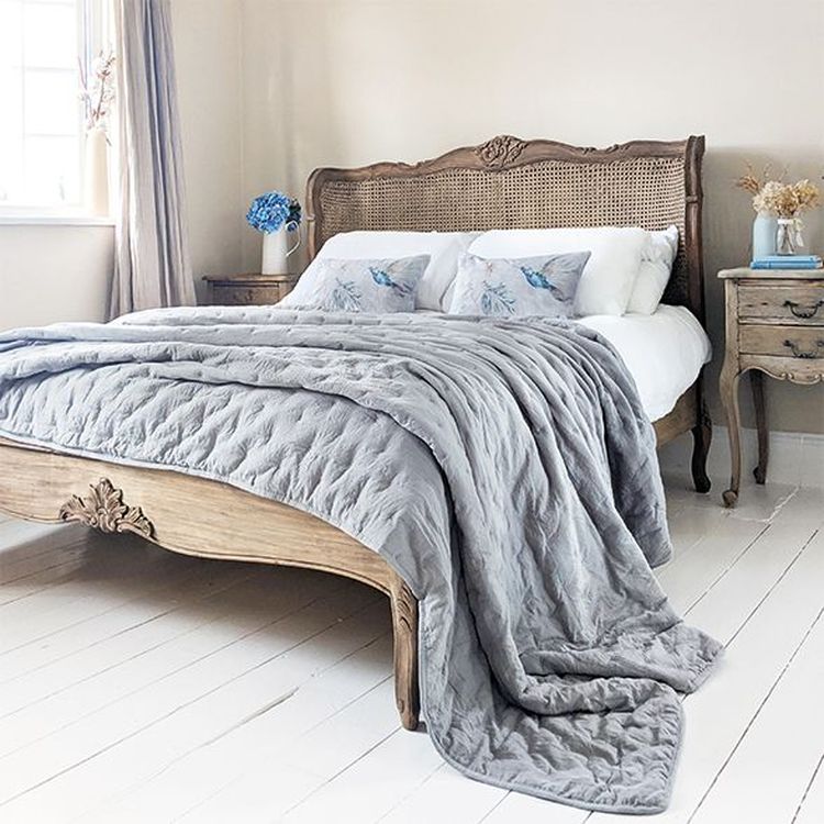 Cozy bedroom with a wooden bed, gray quilt, floral pillows, and vintage nightstands on white wooden floors. Soft natural light from the window.