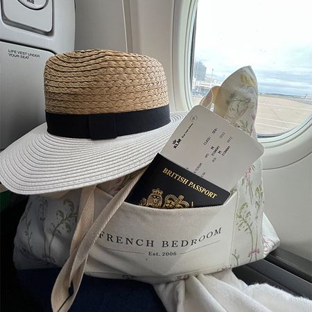 A straw hat, British passport, and boarding pass in a tote bag on an airplane seat by the window.