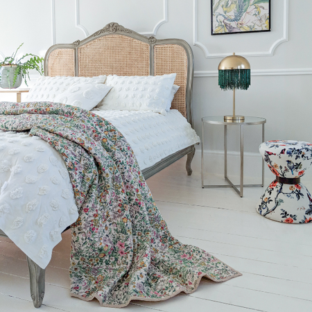 Elegant bedroom with a vintage bed, floral quilt, white pillows, a gold lamp, and a patterned stool on white wooden flooring.