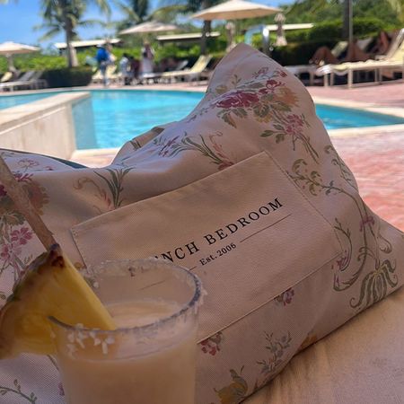 A tropical drink with a pineapple slice sits by a poolside on a floral cushion labeled "French Bedroom." Palm trees and loungers are in the background.