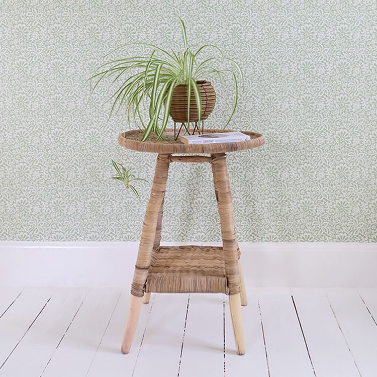 Woven side table with a potted spider plant on top, set against a patterned green wallpaper and white wooden floor.