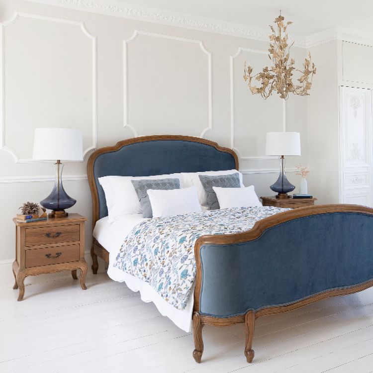 Elegant bedroom with a blue upholstered bed, floral bedding, wooden nightstands, blue lamps, and a gold chandelier against white paneled walls.