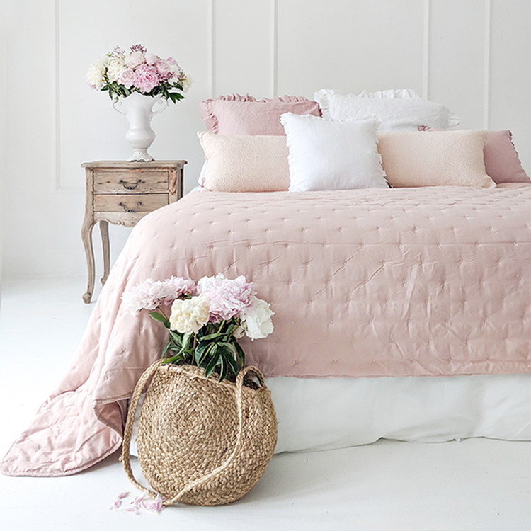 A cozy bedroom with a pink quilted bedspread, assorted pillows, a wooden nightstand with flowers, and a woven basket with peonies.