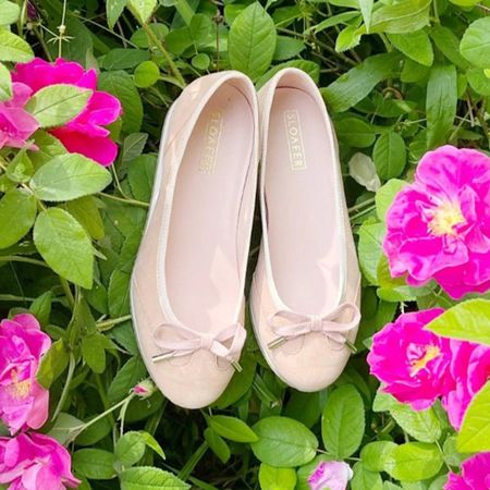 Beige ballet flats with bows placed on lush green leaves, surrounded by vibrant pink flowers.