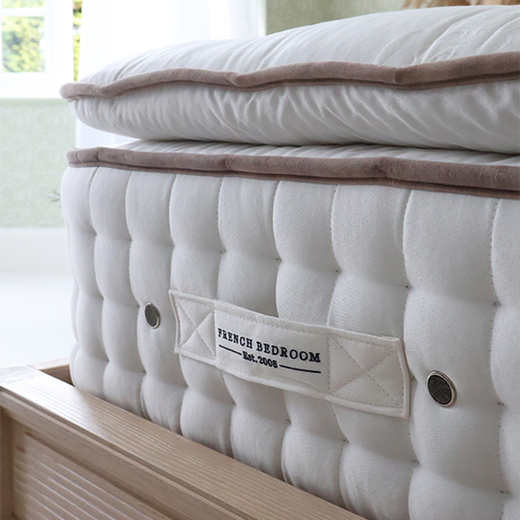 Close-up of a plush, white mattress with a beige trim, featuring a "French Bedroom Est. 2006" label, on a wooden bed frame.