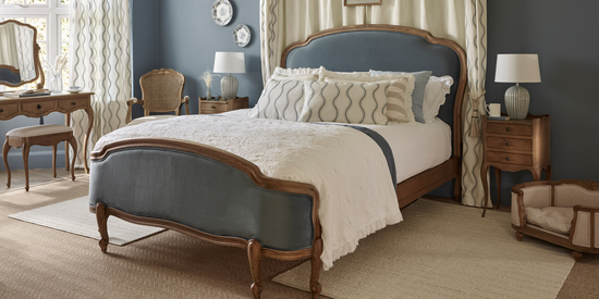 Love Story Blue Velvet Bed made with white textured and ruffled bedding, in a well lit blue walled room surrounded by matching wooden bedside table, upholstered pet bed, rattan chair and dressing table with mirror.