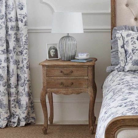 Rustic French bedside table featuring a striped French blue lamp, a photo frame and a tea cup. Besides the bedside table a blue toile curtain hangs and an upholstered bed is dressed with French Toile bed linen.