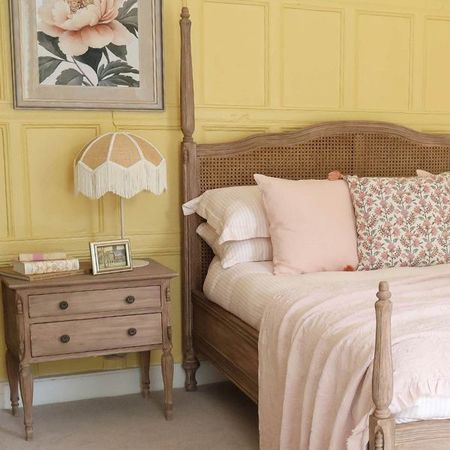 Rattan bed next to a two-drawer French bedside table with a fringe lamp, some books and a picture frame, in front of a yellow wall.
