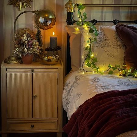 A light wooden bedside in a dark room curated with multiple brass décor items, a plant and a candle atop.
