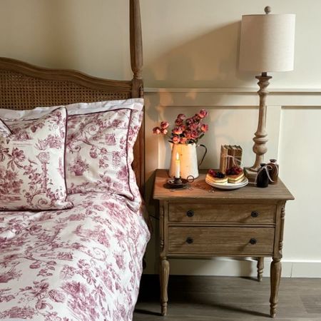 Berry red toile bed linen on a rattan bed next two a wooden bedside table with flowers a candle and pancakes.