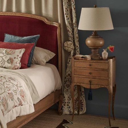 French bedside table with cabriole legs beside a velvet upholstered bed, with a wooden lamp, some books, and a vase with a flower on top.