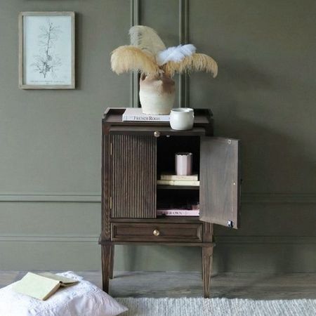 Dark wood bedside table with cupboard doors, a hidden shelf, and a bottom drawer, shows books and candles stored inside while a vase, book and mug sit atop the bedside cabinet.