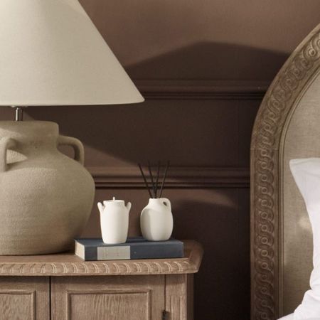 White candle and diffuser placed on a bedside table with a beige table lamp matching a beige upholstered French-style bed