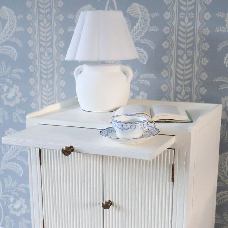 White Bedside table with a white stone effect table lamp and a cup of tea on a pull out shelf.
