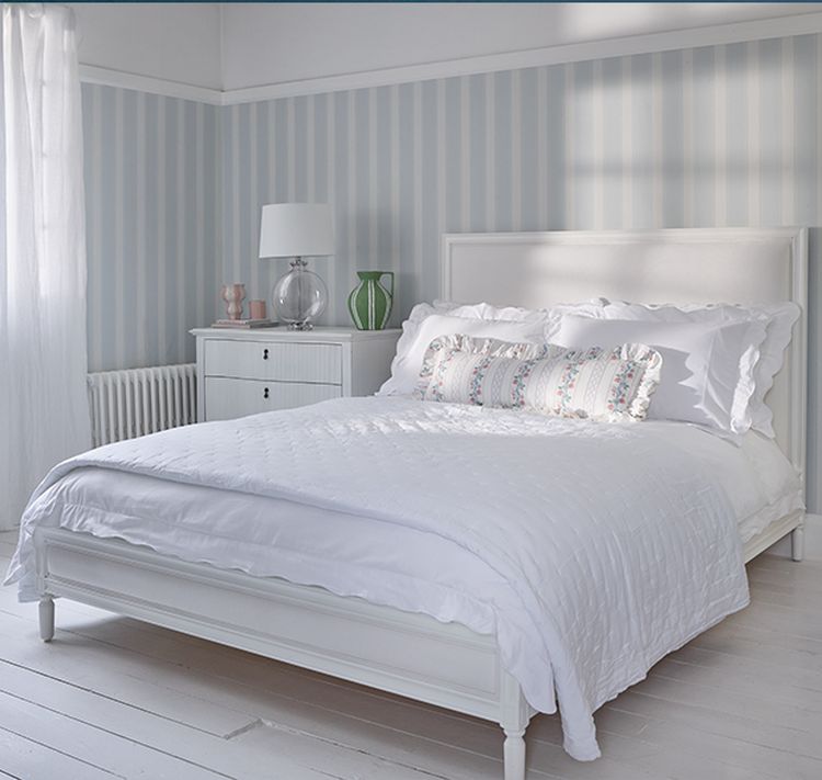 A bright bedroom with a white bed, floral pillows, striped wallpaper, a side table with a lamp, and a vase, all in soft, pastel tones.