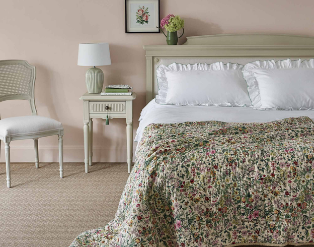 Cozy bedroom with a floral quilt, white pillows, neutral bedside table with a lamp, vase with flowers, and a chair. Neutral tones and soft lighting.
