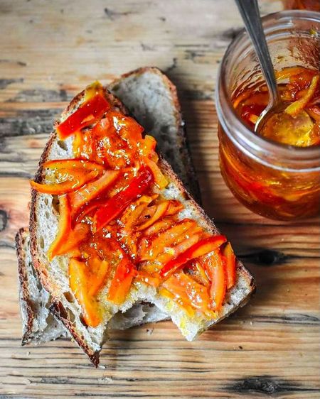 Slice of rustic bread topped with orange marmalade, next to a jar of marmalade with a spoon, on a wooden surface.