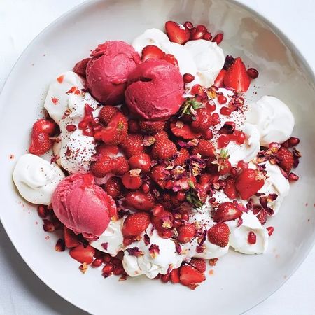 A bowl of whipped cream topped with strawberries, pomegranate seeds, and scoops of pink sorbet, garnished with rose petals.
