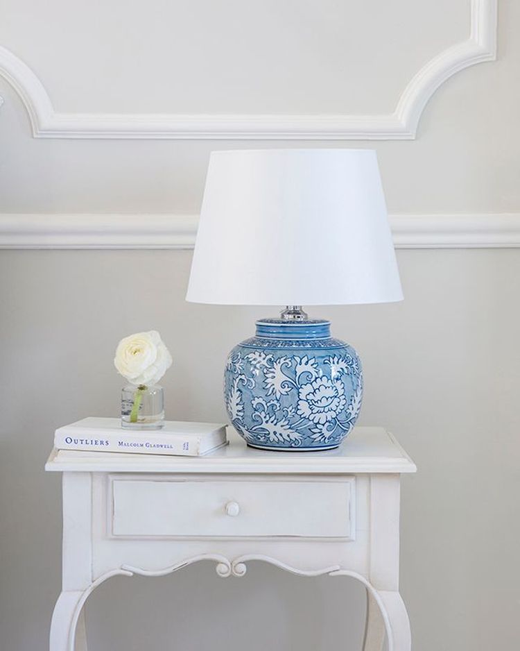 A white lamp with a blue floral base on a vintage white table, next to a book and a small vase with a white rose.
