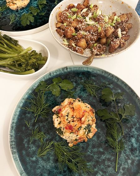 Plated salmon patty with herbs, a bowl of roasted potatoes topped with sesame seeds, and a side of steamed greens.