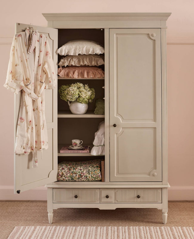 Open wardrobe with a floral robe on the door, shelves holding pillows, a vase of flowers, a cup and book, and a floral blanket at the bottom.