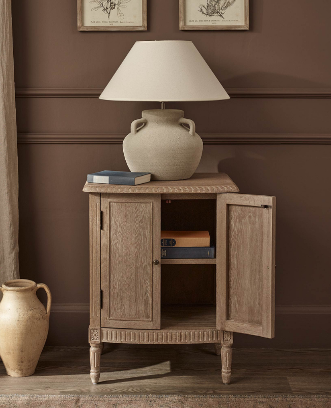 Wooden bedside cabinet with one door open, topped with a beige lamp and books.