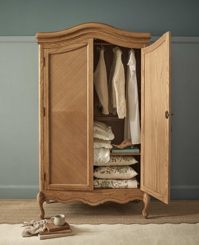 Open wooden wardrobe with hanging clothes and neatly stacked pillows. A stack of books and a mug are on the floor beside it.