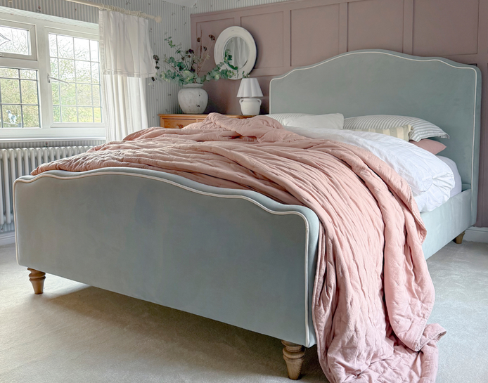 A cozy bedroom with a pastel blue bed, pink duvet, and white pillows. Soft lighting from a window and a vase with flowers decorates the room.