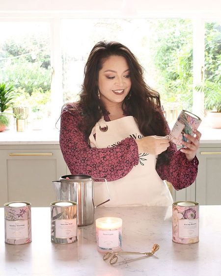 Woman in a floral blouse and apron making candles in a bright kitchen, surrounded by lit candles and greenery outside the window.